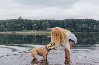 Sarışın, kıvırcık saçlı, labrador köpeğiyle nehirde oynayan genç ve güzel bir kadın..