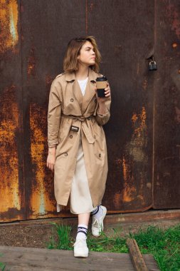 Genç gülümseyen milenyum kadını, sonbahar paltosu giymiş, eski paslı duvarın yanına gitmek için elinde bir fincan kahveyle vahşi saçlarıyla..