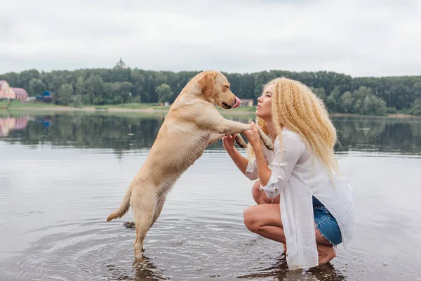 若いです美しいです女性とともにブロンド巻き毛ハグとともに彼女のラブラドール取得犬オンザ川の海岸 — ストック写真