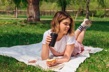Romantik genç bir kadın açık bir kitap, kek ve bir fincan kahveyle parkta çimlere uzanıyor güneşli bir yaz gününde.