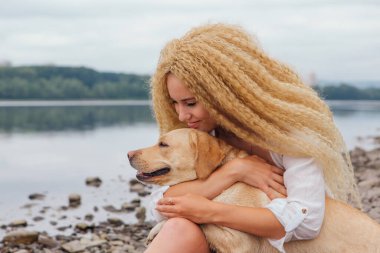 Sarı kıvırcık saçlı genç bir kadın nehir kıyısında Labrador av köpeğiyle sarmaş dolaş.
