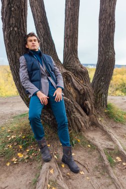 Sarı sonbahar ormanında ağacın yanında duran uzun boylu yakışıklı adam.
