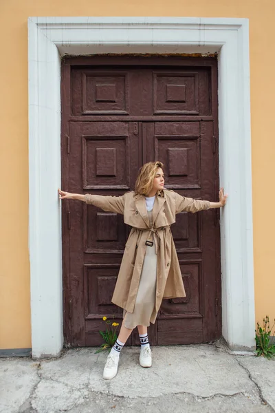 Genç, gülümseyen, vahşi saçlı, sonbahar montu giymiş, eski bir binanın kapısında poz veren bir kadın..