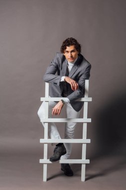 Beyaz balıkçı yaka, beyaz kot pantolon ve gri arka planda beyaz merdivenle poz veren uzun boylu yakışıklı bir adamın moda portresi.