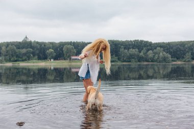 Sarışın, kıvırcık saçlı, labrador köpeğiyle nehirde oynayan genç ve güzel bir kadın..