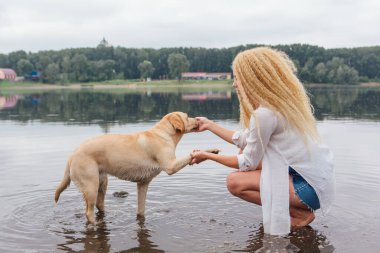 Sarışın, kıvırcık saçlı, labrador köpeğiyle nehirde oynayan genç ve güzel bir kadın..