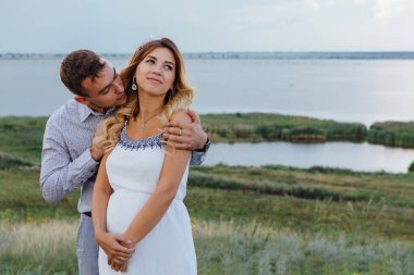 Bebek bekleyen mutlu genç bir çift, yaz yağmurlu bir günde göl kıyısında yürüyorlar. Adam beyaz elbiseli güzel hamile karısına sarılıp öpüyor. Bebeği beklemek, hamilelik konsepti..