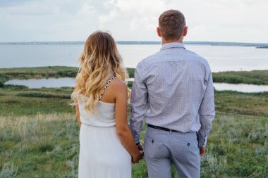 Bebek bekleyen mutlu genç bir çift, yaz yağmurlu bir günde göl kıyısında yürüyorlar. Beyaz elbiseli güzel hamile kadınıyla el ele tutuşan bir adam. Bebeği beklemek, hamilelik konsepti..