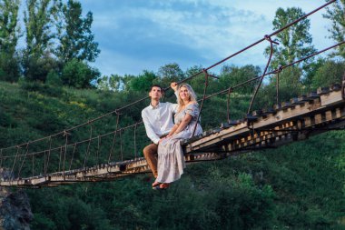 Güzel romantik çift nehrin yukarısındaki asma köprüde oturup sarılıyorlar.