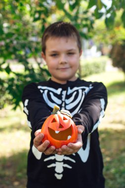 İskelet kostümlü gülümseyen çocuk elinde küçük bir cadılar bayramı balkabağı tutuyor..