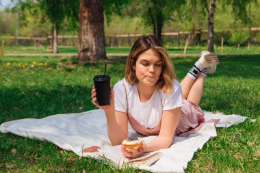 Romantik genç bir kadın açık bir kitap, kek ve bir fincan kahveyle parkta çimlere uzanıyor güneşli bir yaz gününde.
