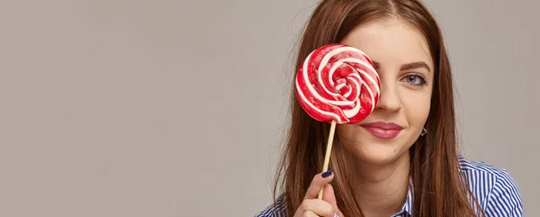 Feliz moderna adolescente mulher sorrindo e se escondendo atrás de um doce pirulito sobre fundo cinza. — Fotografia de Stock