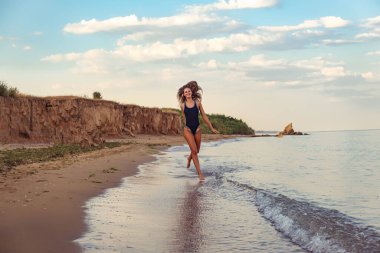 Kumlu sahilde deniz suyunda koşan siyah mayolu mutlu bronzlaşmış genç bir kadın.