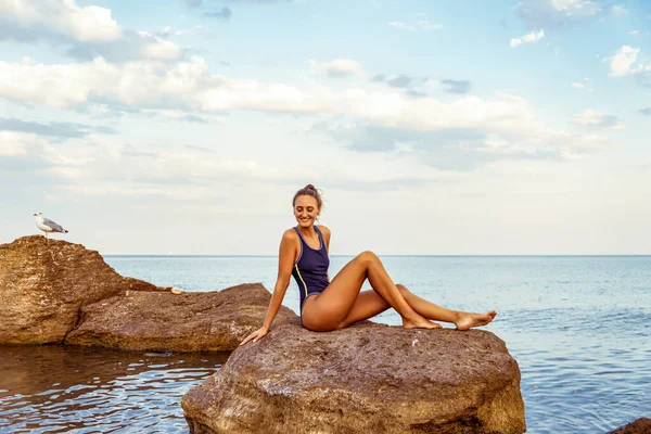 海水中の湿った岩の上に座っている間にスポーティな水着姿で魅力的な日焼けした若い女性 — ストック写真
