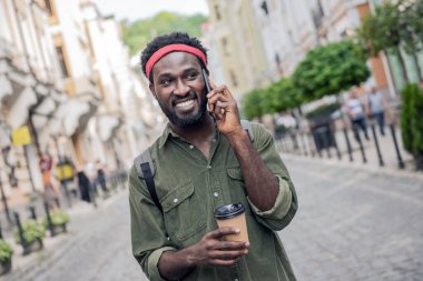 Genç siyah adam gülüyor ve kahve içiyor. Sırt çantasıyla köprüde yürürken telefonda konuşuyor.