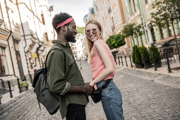 街の通りを歩いているときに暗い肌の男とブロンドの女性がお互いに手を取り合って笑っている若い陽気なカップル — ストック写真