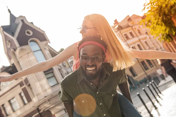 黒人男性と金髪の女性が笑っている若い陽気なカップル 街を歩いている間に腕を伸ばして男の後ろに座っている女の子 — ストック写真