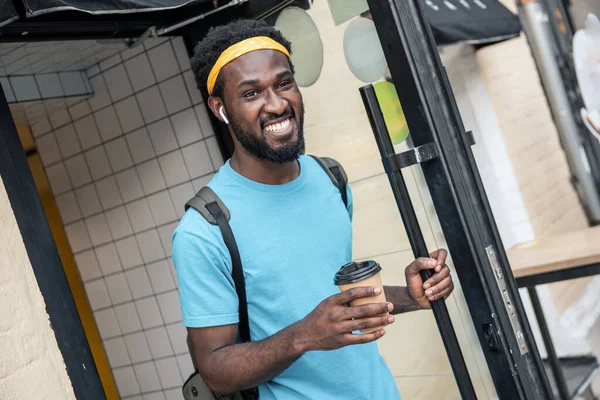 バックパックを持って橋の通りを歩いている間にオープンドアを保持してカフェから出てくる若い黒人男性飲料コーヒー — ストック写真