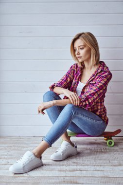 Kaykayın üzerinde oturan ve arka planda kameraya gülümseyen neşeli güzel sarışın kızın portresi.