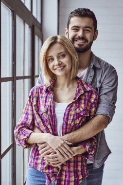 Genç ve neşeli bir çiftin portresi. Gülümseyen, kameraya bakan ve birbirlerine sarılan ve pencereden dışarı bakan.