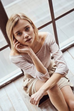 Genç ve güzel bir kızın portresi büyük panoramik pencerenin yanında oturuyor ve kameraya gülümsüyor.