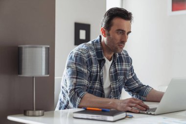 Genç bir erkek işadamı dizüstü bilgisayarla çalışırken mutfaktaki masada oturup bir şeyler düşünüyor.