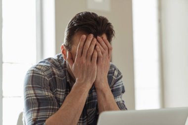 Genç erkek işadamı dizüstü bilgisayarla çalışırken mutfakta masada oturuyor ve yorgun bir şekilde elleri ile yüzünü kapatıyor.