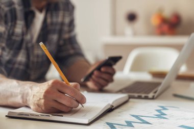 Genç bir iş adamının, mutfaktaki bir masada dizüstü bilgisayarla uzaktan çekilmiş bir fotoğrafı. Kalemle deftere yazıyor ve diğer el de telefonu kullanıyor..