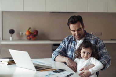 Uzaktan kumandalı bir baba mutfaktaki evinde bir masada oturuyor ve küçük kızı kucağında oturmuş ilgi bekliyor.