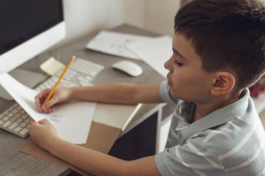 Küçük çocuk odasında bilgisayarın yanında oturuyor ve okul için ödevlerini hazırlıyor.
