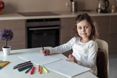 Küçük mutlu kız evde, mutfakta bir masada oturmuş renkli kalemler ve kalemlerle bir resim çiziyor.