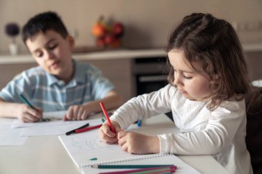 Küçük mutlu erkek ve kız kardeş mutfaktaki bir masada oturup renkli kalemler ve kalemlerle bir resim çiziyorlar.