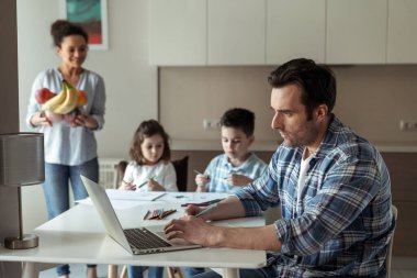 Annem evde bilgisayar ve belgelerle çalışırken babaya meyve ikram eder. Mutfaktaki masada otururlar. Kızları ve oğulları renkli kalemler ve kalemlerle çizerler.