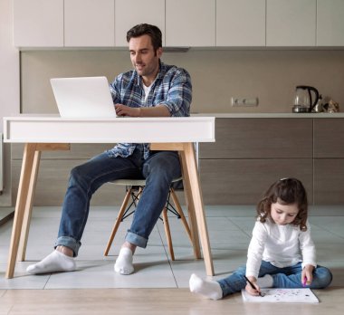Babam evde bilgisayar ve belgelerle çalışıyor. Mutfaktaki bir masada oturuyor. Ve yere renkli kalemler ve kalemlerle resim çizen küçük kızı.