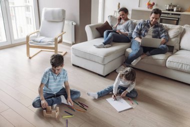 Küçük oğlan ve kız, evde çalışan anne ve babalarının arka planında dizüstü bilgisayar ve tabletle oturup oyun oynuyor ve resim çiziyorlar.