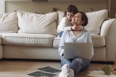 Evde dizüstü bilgisayar ve belgelerle çalışırken kanepenin yanında oturan başarılı serbest çalışan genç kadın annesinin boynuna sarılıyor.