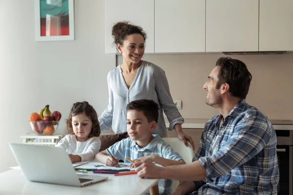 Mom helps little daughter and son draw while father works at home with computer and documents sitting at table in kitchen and smiling looking towards his wife