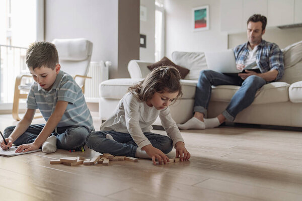 Маленький мальчик и девочка играют и рисуют, сидя на полу на фоне своего отца, который работает дома, сидя на диване с ноутбуком