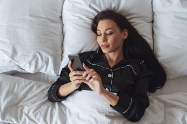 Genç ve güzel bir kadın yatakta uyuduktan sonra uyandı ve yatak odasındaki battaniyenin altında pijamalarıyla telefonu kullanıyor..