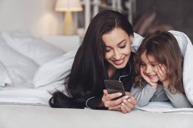 İyi geceler ya da günaydın. Genç, güzel anne ve küçük kızı evde yatakta yatarken ve kameraya gülümserken telefonu kullanıyorlar..