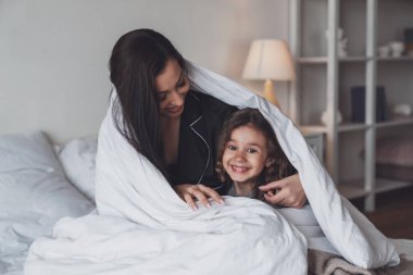 İyi geceler ya da günaydın. Mutlu genç anne ve küçük kızı battaniyeyle örtülmüş ve kameraya gülümsüyor..