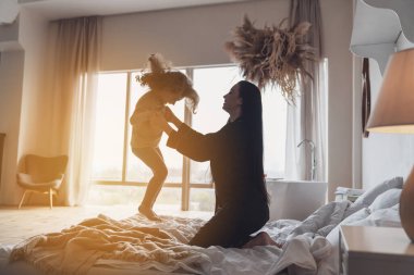 İyi sabahlar. Mutlu genç anne ve küçük kızı evdeki yatak odasında büyük bir pencerenin arkasında zıplıyorlar..