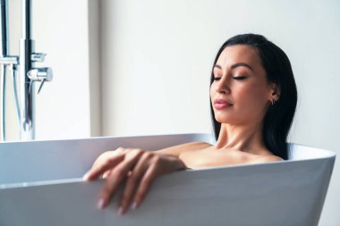 Genç ve güzel bir kadın büyük bir pencerenin arkasında evdeki banyoda banyo yaparken rahatlıyor..