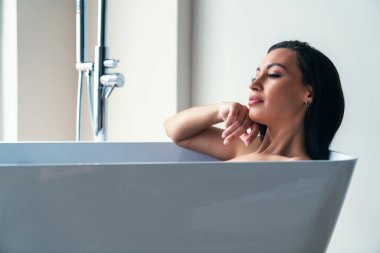 Genç ve güzel bir kadın büyük bir pencerenin arkasında evdeki banyoda banyo yaparken rahatlıyor..