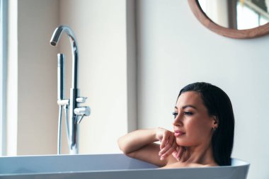 Genç ve güzel bir kadın büyük bir pencerenin arkasında evdeki banyoda banyo yaparken rahatlıyor..