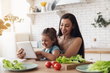 Küçük mutlu bir kız annesinin öğle yemeği için salata hazırlamasına yardım ediyor. Mutfakta bir masada oturuyor ve elektronik tablet kullanıyor..