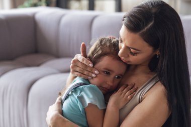 Genç bir anne, evdeki oturma odasında ona yaslanmış ağlayan küçük bebeğini sakinleştirir..