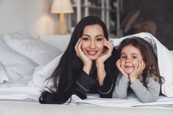 Good night or good morning. Happy young mother and her little daughter lying on the bed at home, covered with a blanket and smiling at the camera.