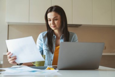 Genç başarılı iş kadını evde çalışırken masada oturmuş, dizüstü bilgisayar kullanıyor ve kahve içiyor..