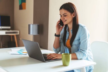 Genç başarılı iş kadını evde çalışırken masada oturmuş, dizüstü bilgisayar kullanıyor ve kahve içiyor..
